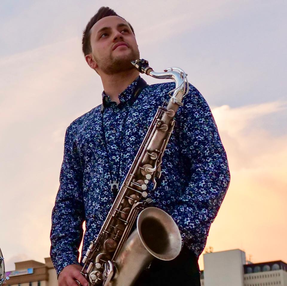 A person holding a saxophone looking off into the distance with a bright sky background